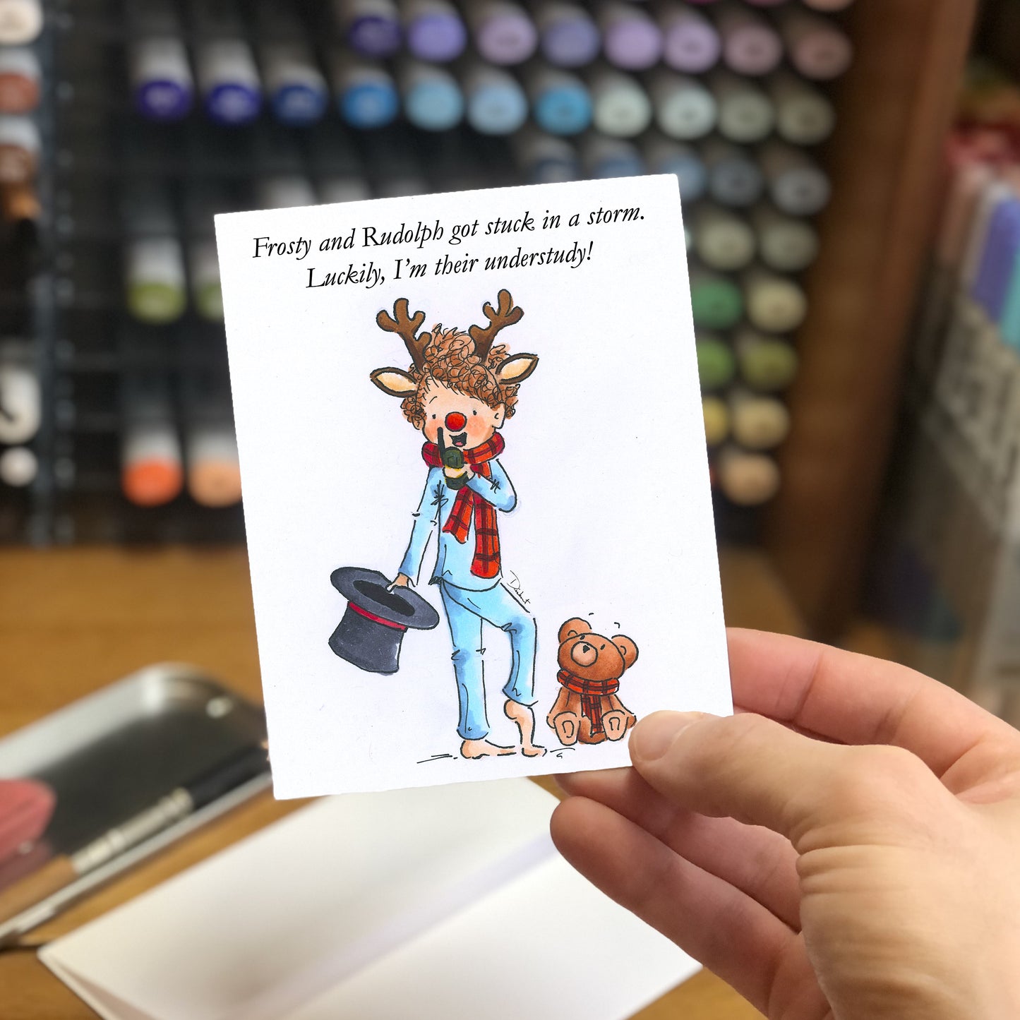 Hand holding a card with a cartoon reindeer and teddy bear, with text about Frosty and Rudolph, in front of a color swatch background.